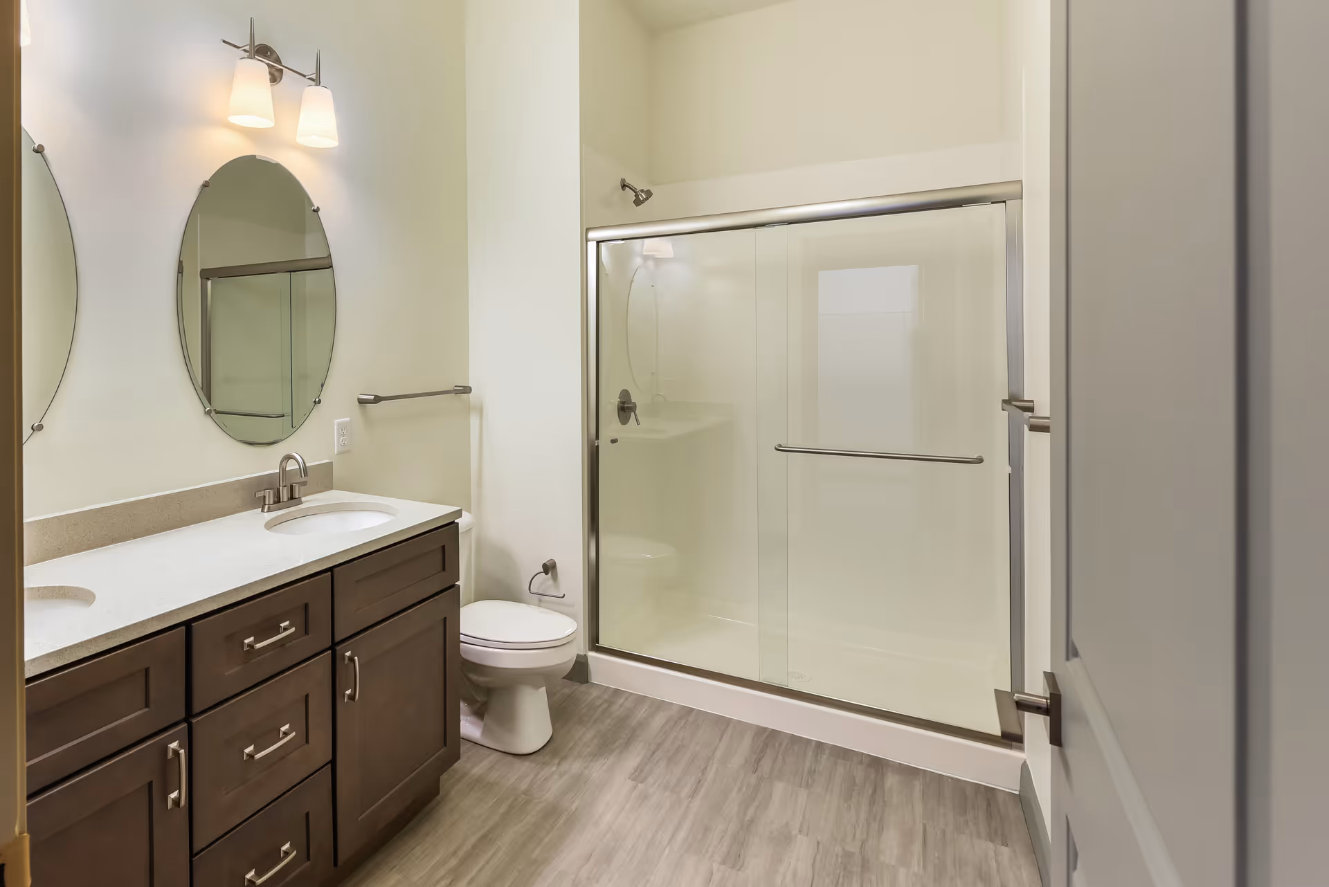 A modern bathroom featuring a double sink vanity with dark wood cabinets and a white countertop. Above the sinks are two oval mirrors and a light fixture with two lamps. To the right is a toilet and a glass-enclosed shower with sliding doors. The floor has a light wood-like finish.