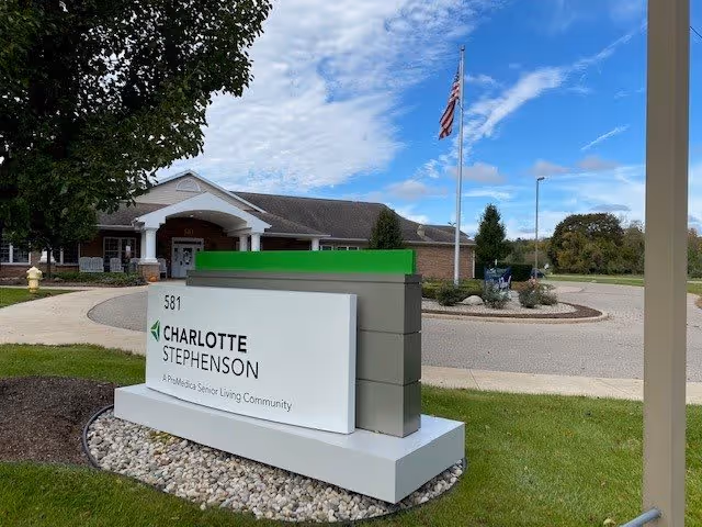 Exterior view of Charlotte Stephenson A ProMedica Senior Living Community building with a sign in the foreground displaying the facility name and address 581. The building has a brick facade, white columns, and an American flag on a flagpole. The sky is partly cloudy with blue patches.