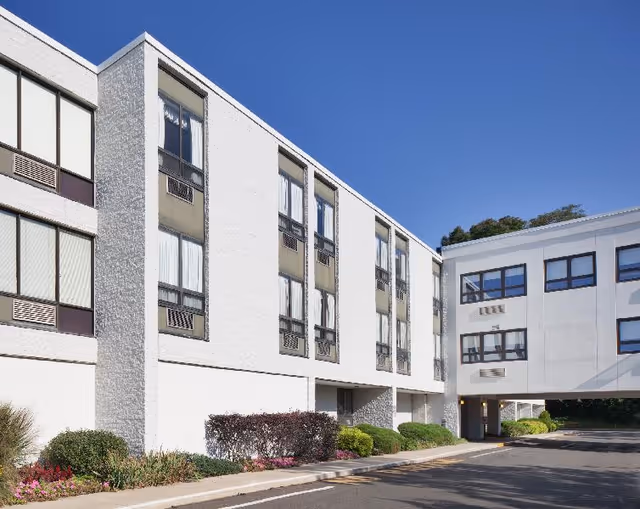 White multi-story building with rows of windows, a covered drive-through entrance and landscaped shrubs under a clear blue sky.