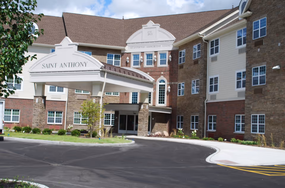 Exterior view of the Saint Anthony senior living facility building with a covered entrance, multiple windows, and a paved driveway in front. The building has a mix of brick and stone facade with a brown roof under a partly cloudy sky.