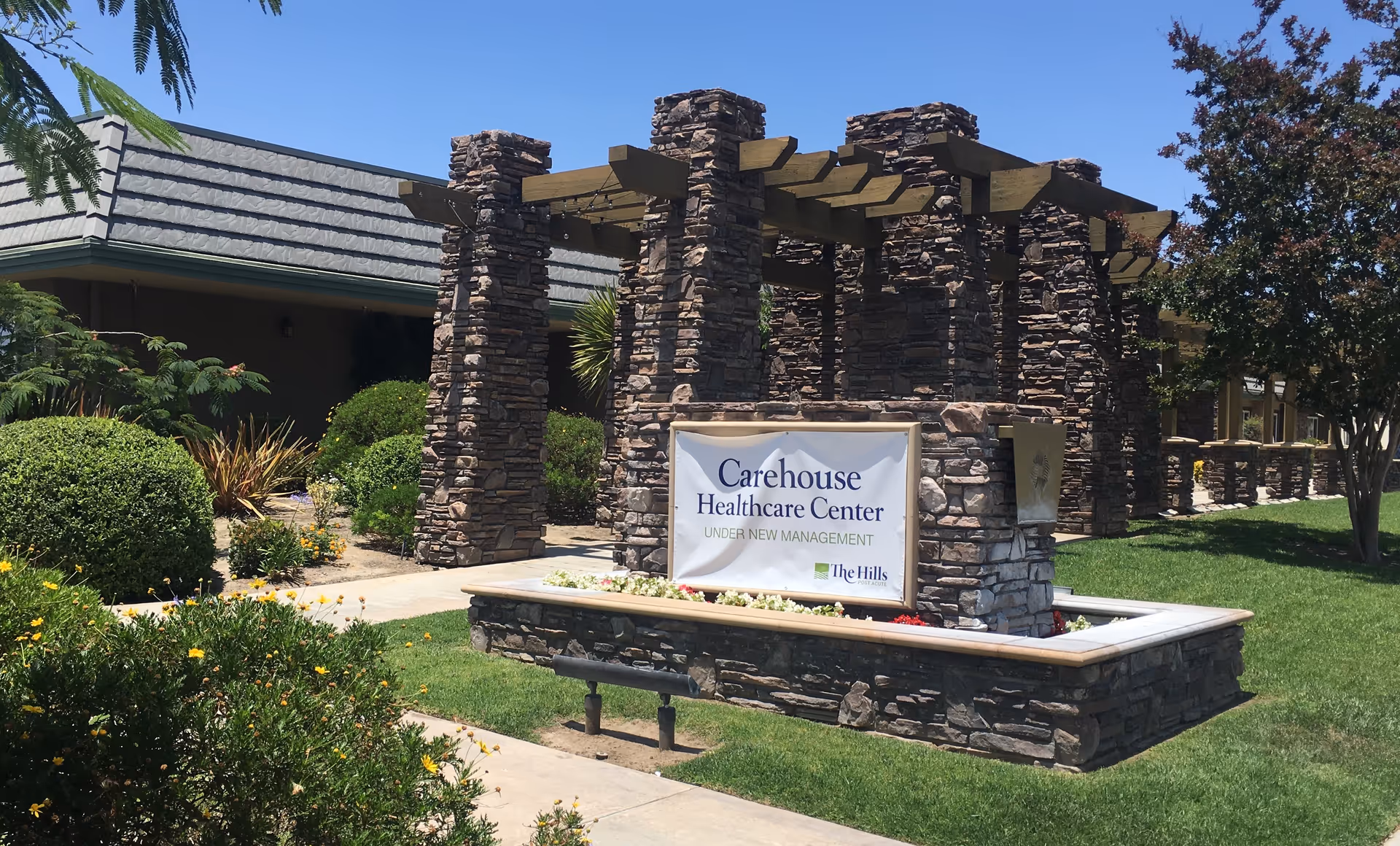 Exterior view of Carehouse Healthcare Center featuring a stone pergola structure and a sign that reads 'Carehouse Healthcare Center UNDER NEW MANAGEMENT The Hills' surrounded by well-maintained greenery and a clear blue sky.