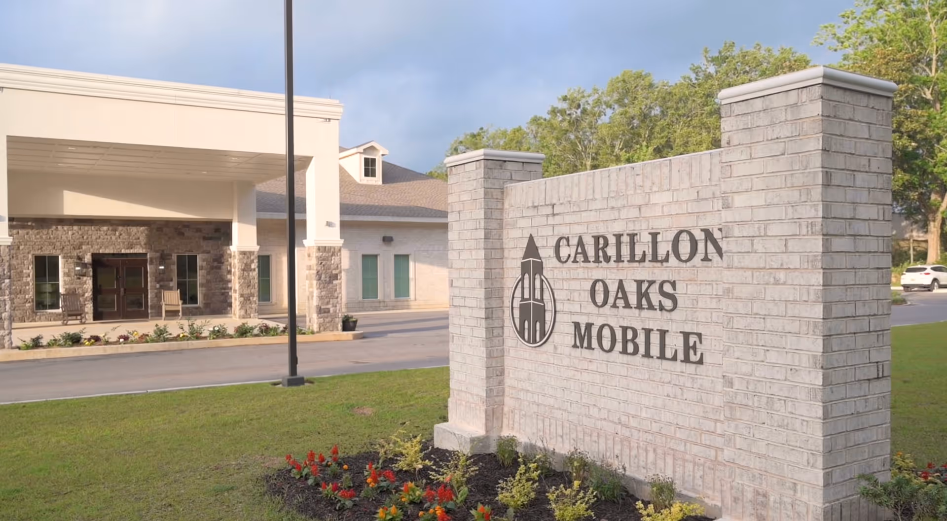 Exterior view of Carillon Oaks Mobile senior living facility showing a large brick sign with the facility name and logo, with the building entrance and landscaping visible in the background.