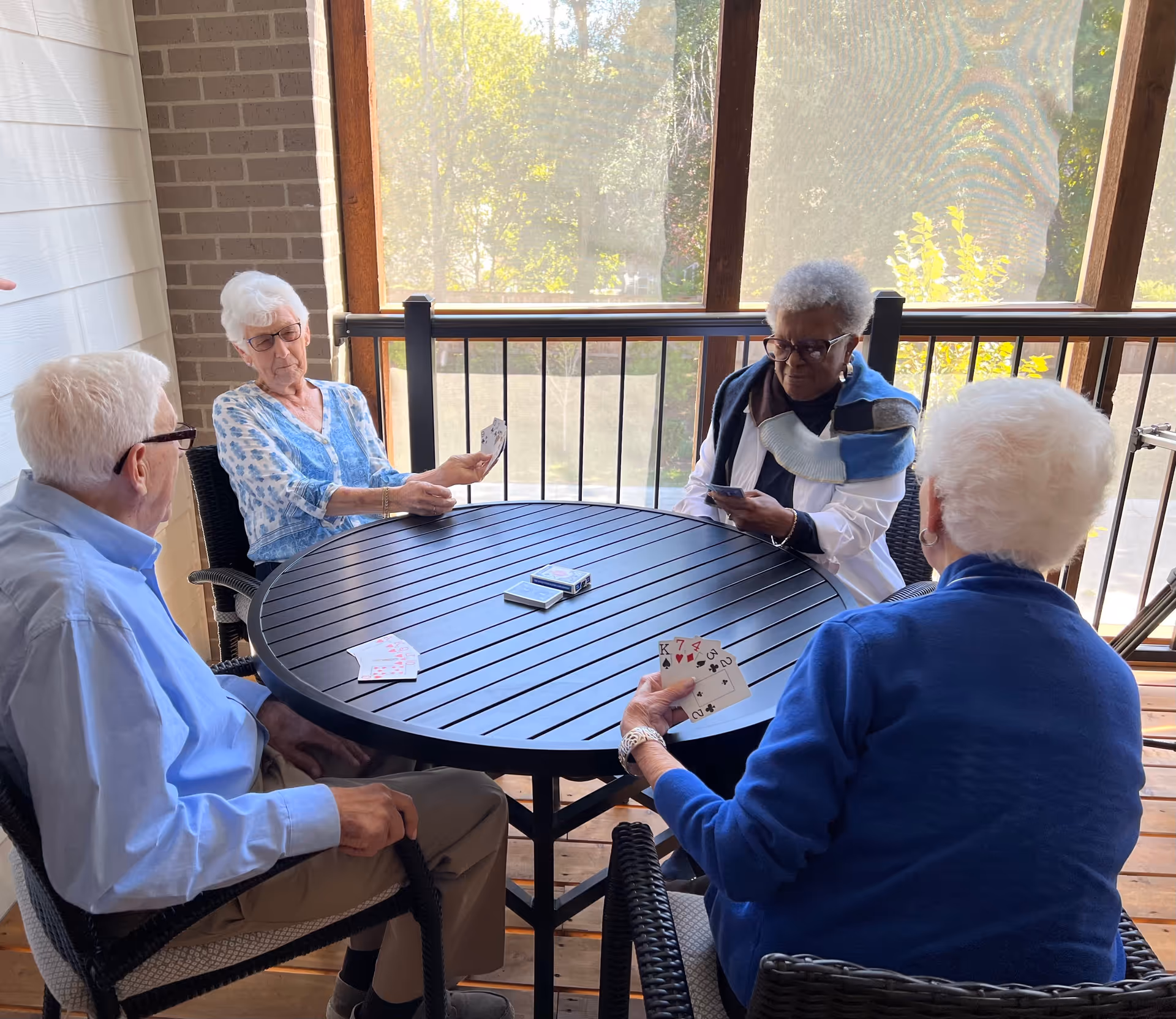 Four elderly people sitting around a round black table on a screened porch, playing cards. The porch has wooden flooring and a railing with a view of trees outside.