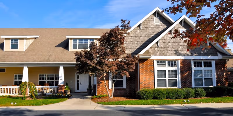 Front exterior view of a residential-style assisted living facility building with a combination of brick and siding, a covered porch with white columns, several windows, and a tree with autumn-colored leaves in front.