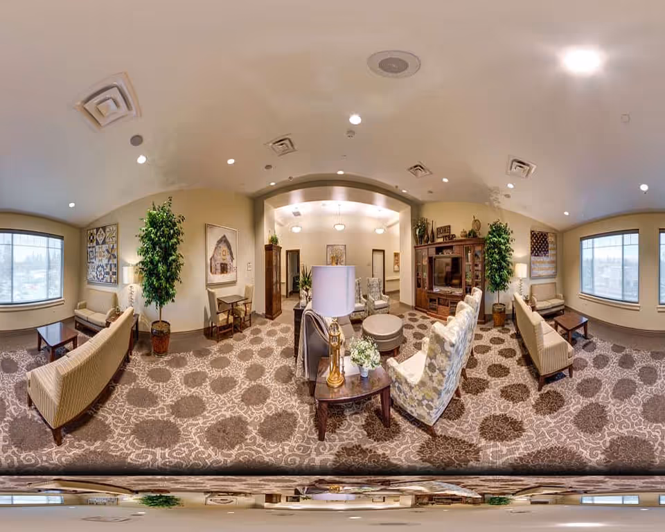 A spacious and well-lit assisted living facility common area with patterned carpet, multiple seating arrangements including armchairs and sofas, decorative plants, framed artwork on the walls, large windows letting in natural light, and a wooden entertainment center with a television.