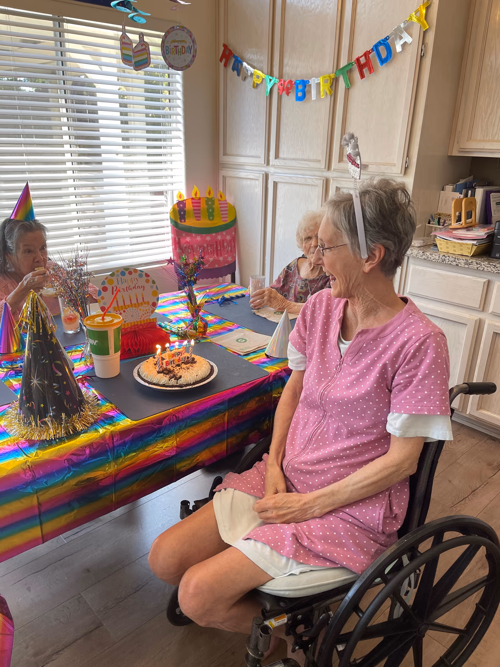 Three elderly women celebrating a birthday in a decorated room. One woman in a wheelchair wearing a pink polka dot dress and a birthday headband is smiling at a birthday cake with lit candles on a table covered with a colorful rainbow tablecloth. The table also has party hats, a drink cup, and birthday decorations. A 'Happy Birthday' banner is hanging on the wall behind them.