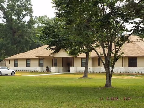 Single-story building with a beige exterior and brown roof surrounded by green grass and trees, with a white car parked on the left side.