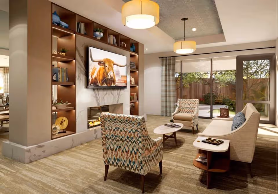 A bright living room with patterned armchairs and a sofa arranged around a coffee table, a TV mounted above a fireplace flanked by built-in shelves, and sliding glass doors to an outdoor patio.