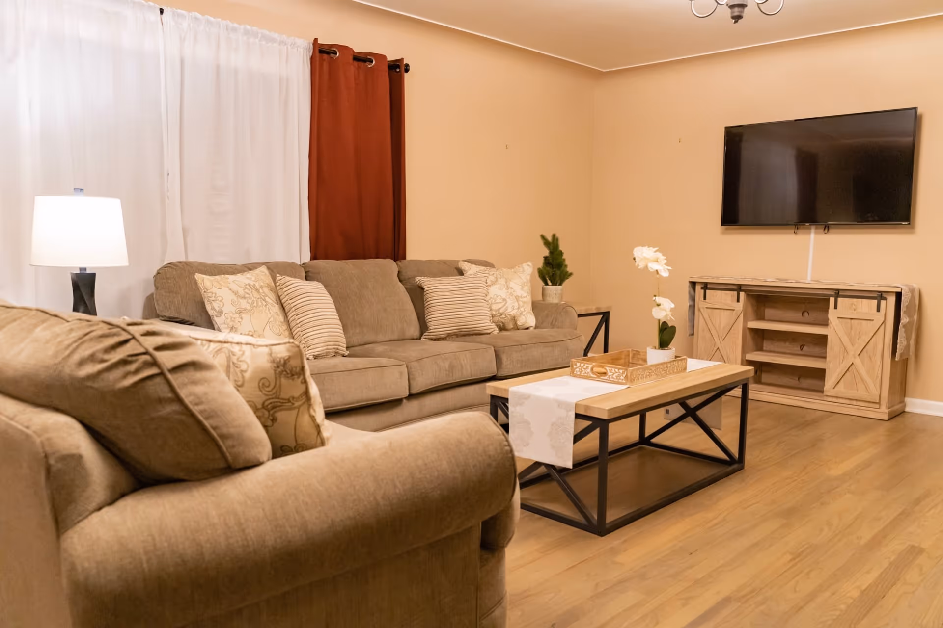 Cozy living room with beige sofas, a coffee table, a wall-mounted TV, and a lamp by a curtained window.