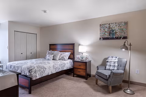 Bright bedroom with a wooden bed, patterned bedding, a nightstand and lamp, and a gray armchair beneath wall art.