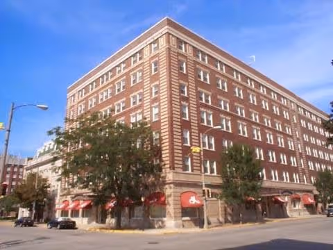 Corner view of a multi-story brick residential building with red awnings and street trees under a blue sky.