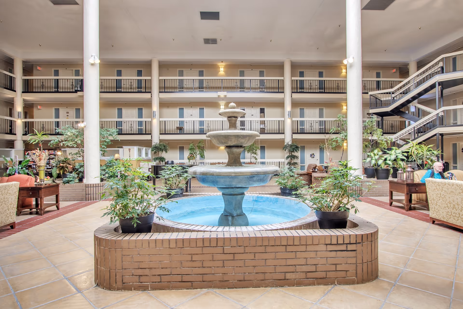 Indoor atrium area of a senior living facility with a large three-tiered water fountain in the center surrounded by potted plants. The space is open with tiled floors, multiple seating areas with chairs and tables, and balconies with doors on three levels. A staircase is visible on the right side, and soft lighting illuminates the area.