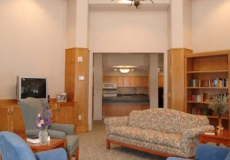 A cozy senior living common room with upholstered sofas and armchairs, a bookshelf, a TV, and a view into a kitchen area.