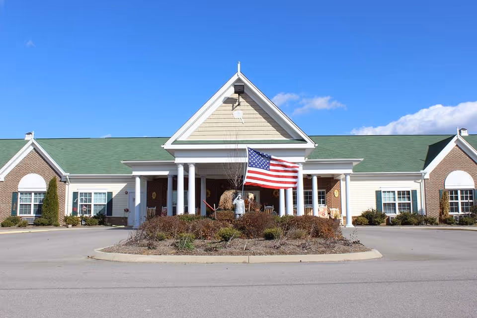 Front exterior view of Meadow View Senior Living facility with a green roof, white columns at the entrance, an American flag in the center, and clear blue sky in the background.