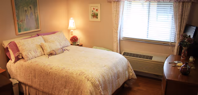 A cozy bedroom with a neatly made bed covered in a floral bedspread and multiple pillows. There is a bedside table with a lamp and a small flower arrangement. A window with blinds and floral curtains lets in natural light. A wooden dresser with decorative items is positioned near the window. The walls are painted a warm beige color and decorated with framed artwork.