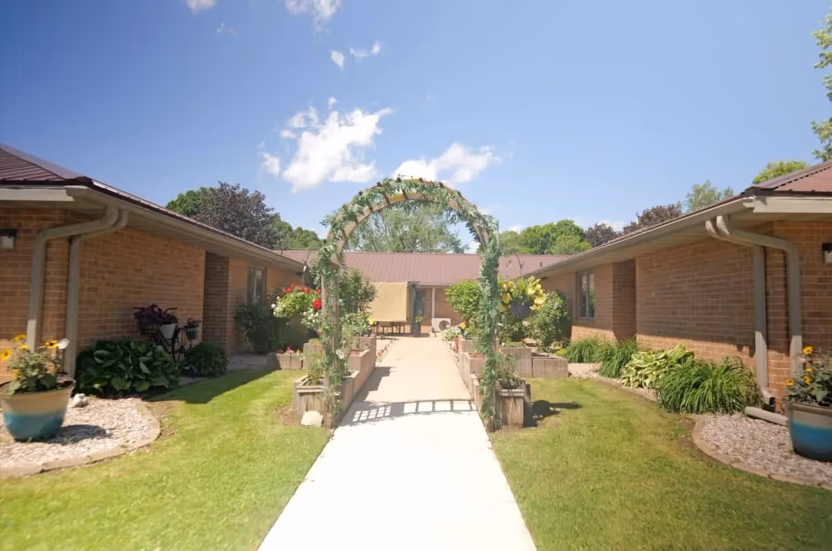 A sunny courtyard between single-story brick buildings with a central walkway and a decorative plant-covered archway.