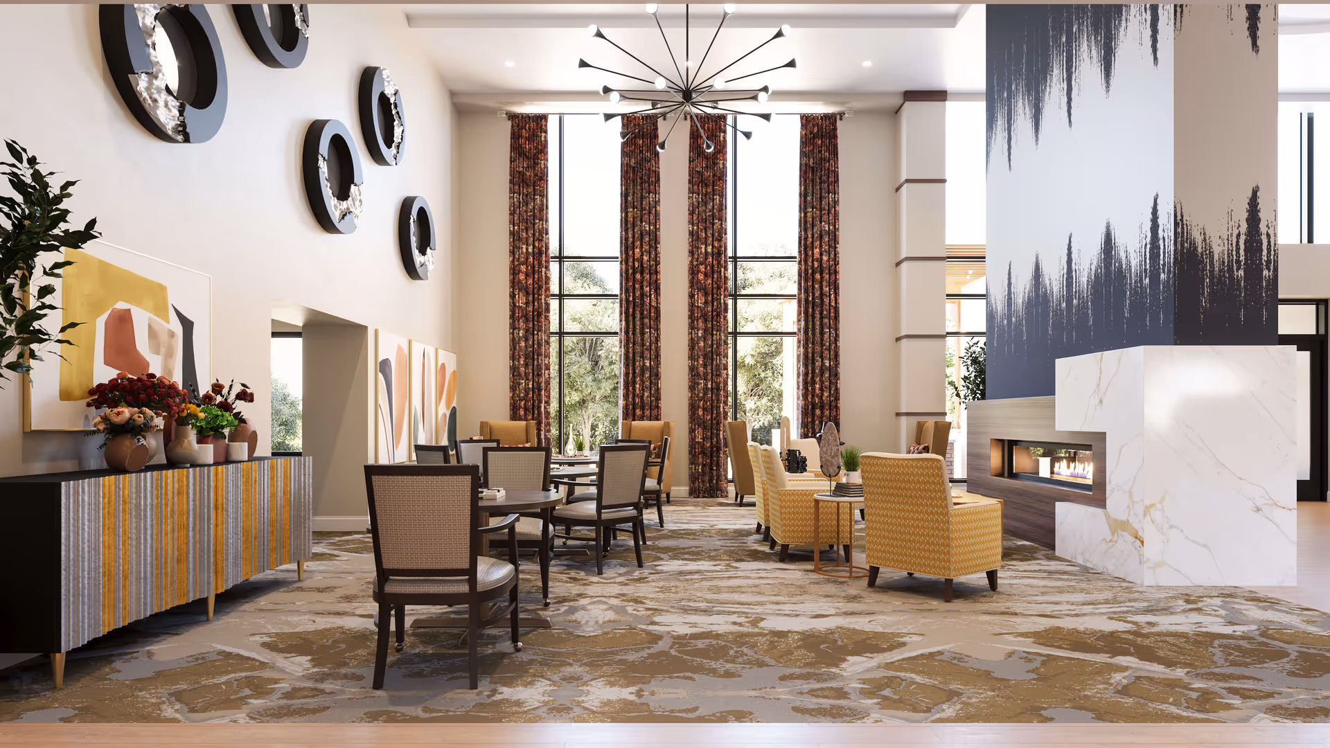 A spacious and well-lit common area with high ceilings and large windows covered by floral curtains. The room features several round tables with chairs, a seating area with yellow patterned armchairs around a modern fireplace with a marble finish, and decorative wall art including circular sculptures and abstract paintings. A contemporary chandelier hangs from the ceiling, and a sideboard with vases and flowers is positioned against one wall.