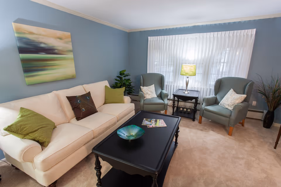 Light-filled living room with a white sofa, two green armchairs, a black coffee table, and a lamp in front of a large window.