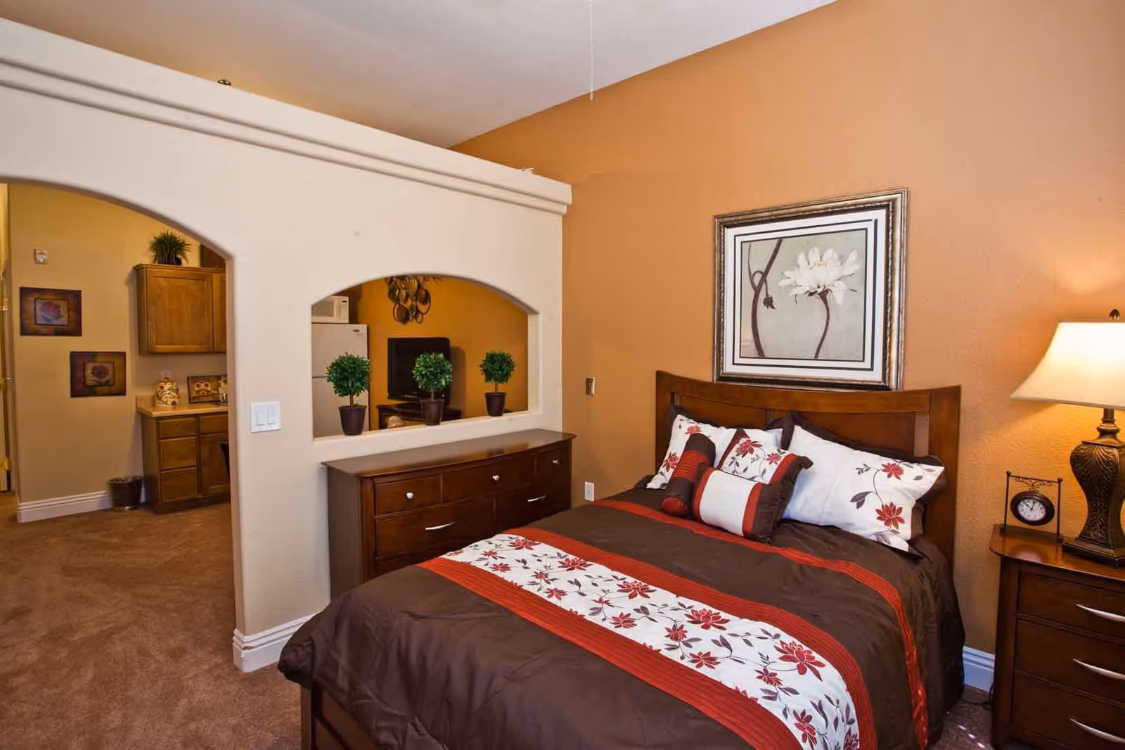 A cozy bedroom with a bed dressed in brown, white, and red floral bedding. There is a wooden headboard and a framed floral artwork hanging above it. To the right of the bed is a nightstand with a lamp and a small clock. A dresser with three small potted plants is positioned against a half wall with two arched openings, which looks into a kitchen area with wooden cabinets and a refrigerator.