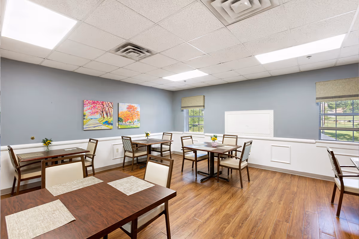 Dining room with several wooden tables and chairs, placemats, small flower centerpieces, and colorful paintings on a gray wall.