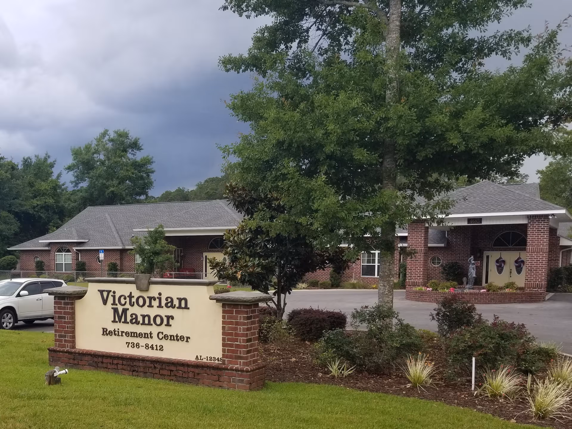 Front view of Victorian Manor retirement center with a brick building, entrance driveway, and a large sign on the lawn.