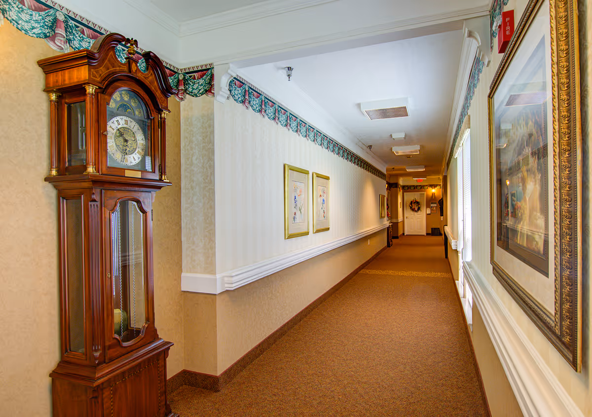 Carpeted interior hallway of a senior living facility with a large grandfather clock, framed artwork, and decorative wall trim.