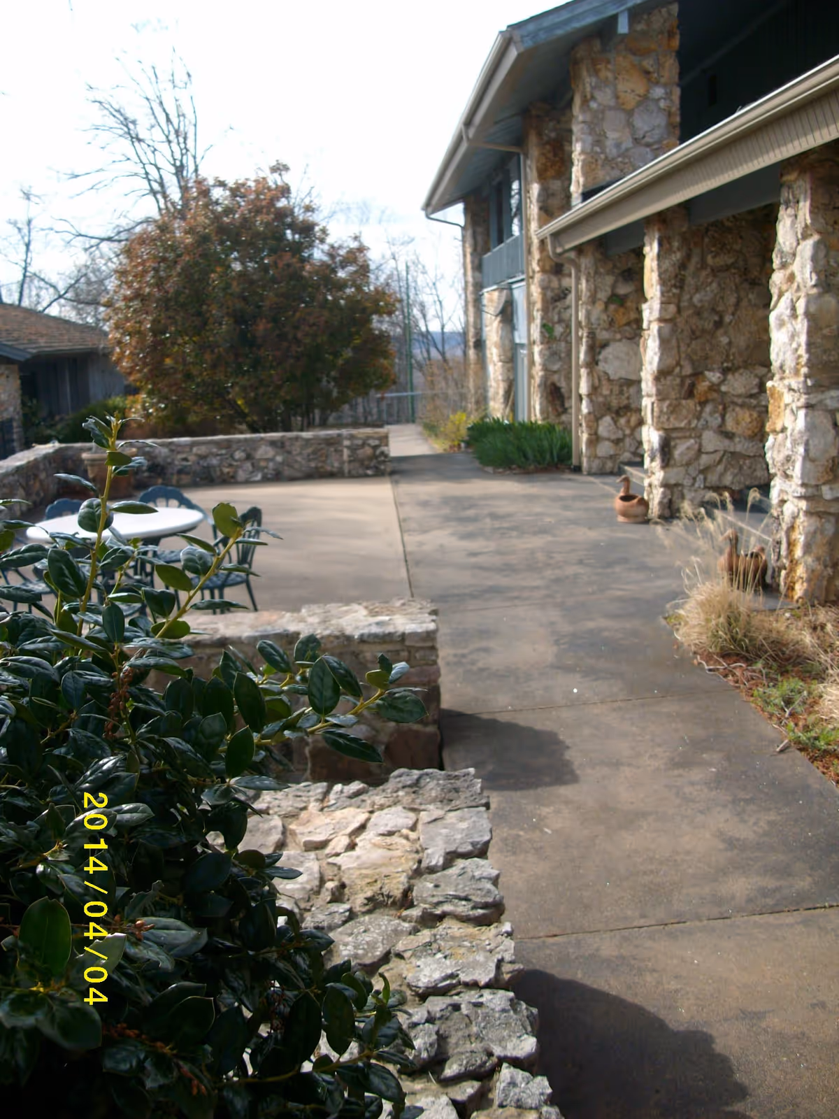 Outdoor patio area with stone walls and columns, a concrete walkway, green plants, and a round table with chairs. The building has a rustic stone exterior and there are trees in the background.