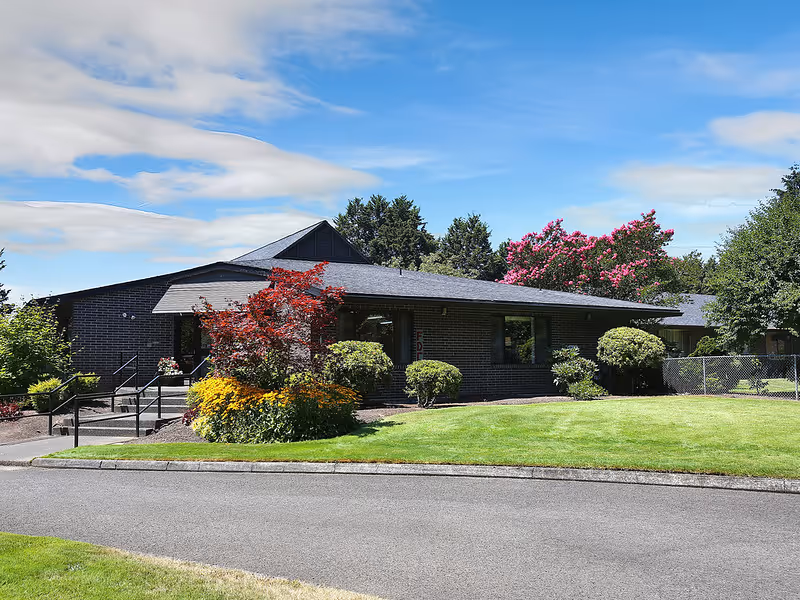 Single-story brick building with a dark roof surrounded by well-maintained green lawn, shrubs, and flowering trees under a partly cloudy blue sky.