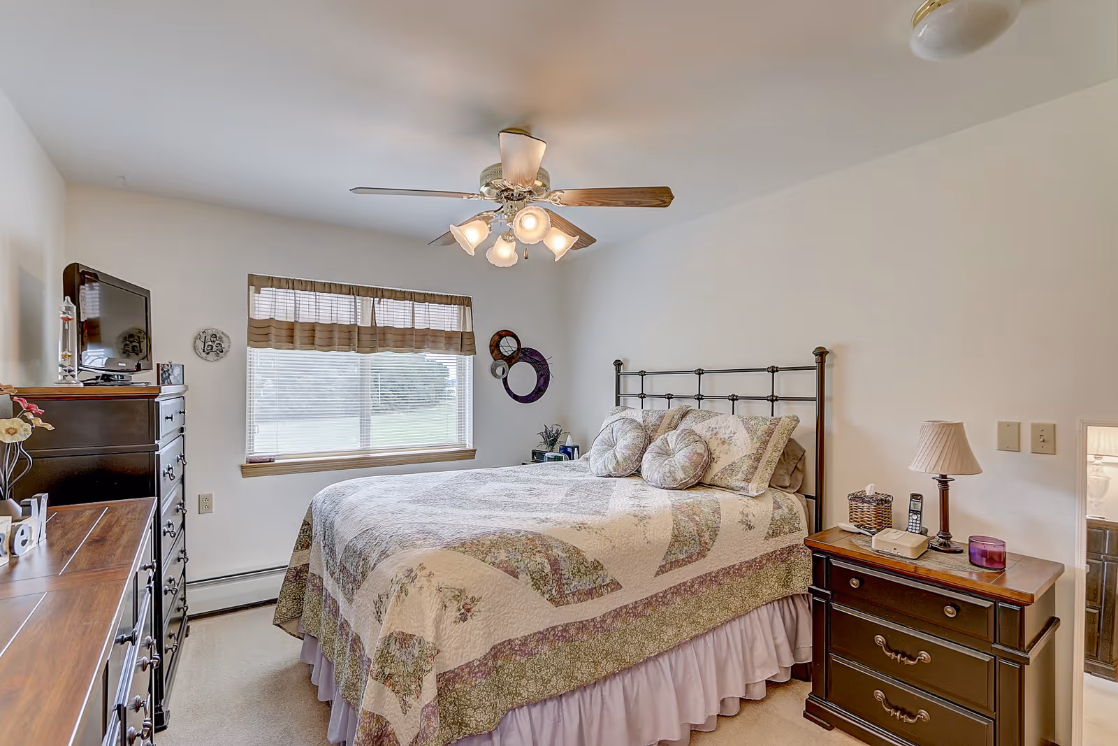 A cozy bedroom with a large bed covered in a floral quilt and matching pillows. The room features a ceiling fan with lights, a window with blinds and a valance, a dark wooden dresser with a TV on top, and a matching nightstand with a lamp, phone, and other small items. The walls are painted white and the room has a soft carpeted floor.