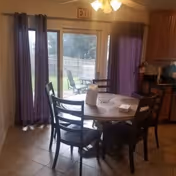 Dining area with a round wooden table surrounded by four chairs. The table has a paper towel roll, a small basket, and a white item on it. Behind the table are sliding glass doors with purple curtains, leading to an outdoor patio with chairs and a wooden fence. The room has tiled flooring and a ceiling fan with lights. Kitchen cabinets and a fire extinguisher are visible on the right side.