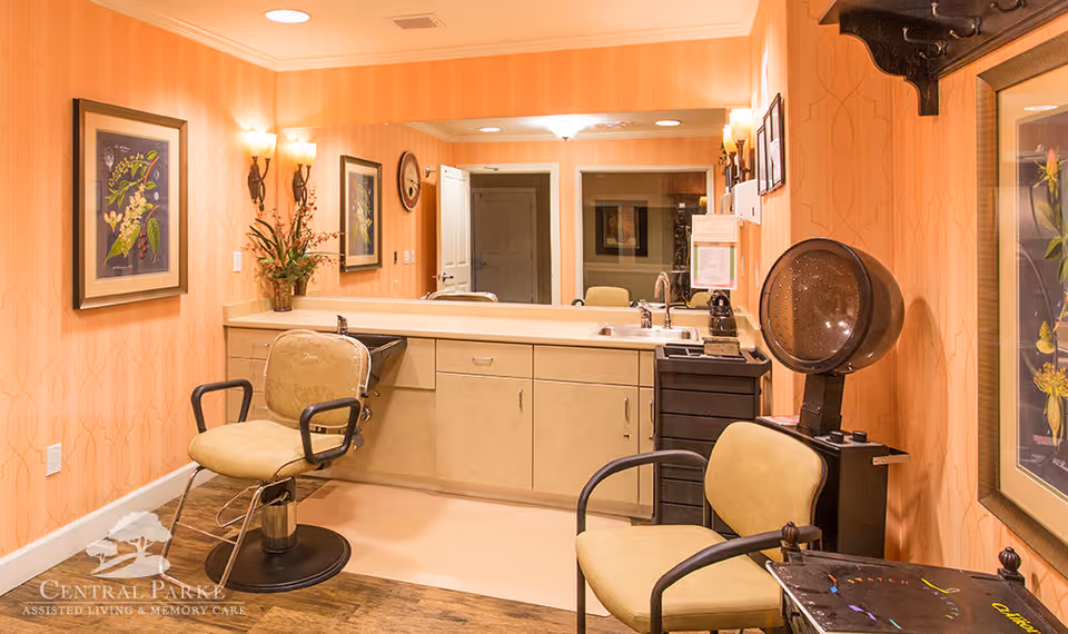 Interior view of a salon area in an assisted living facility with two salon chairs, a hair dryer, a large mirror, framed botanical artwork on peach-colored walls, and a countertop with a sink and cabinets underneath.