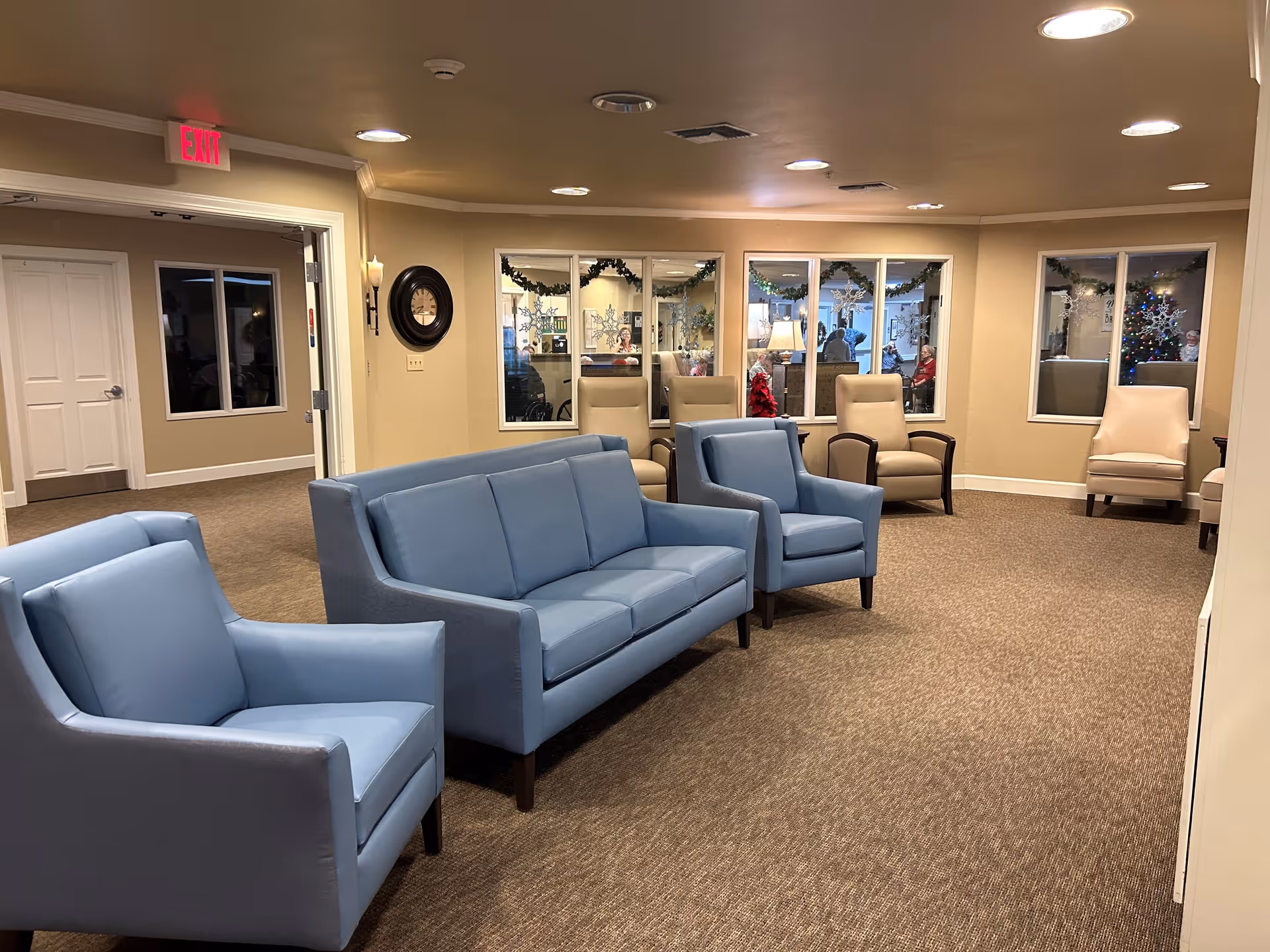 A cozy seating area in a senior living facility with light blue armchairs and a matching sofa arranged on a carpeted floor. Beige walls and ceiling lights illuminate the room. Several beige armchairs are placed near windows decorated with holiday garlands and snowflake decals. A Christmas tree is visible through one of the windows, and an exit sign is above a doorway on the left.