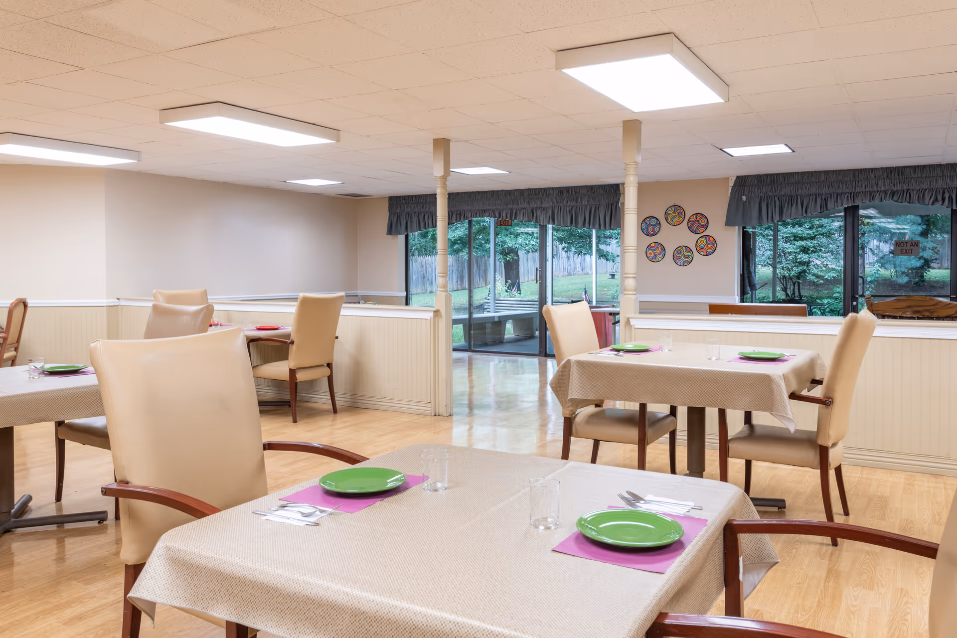 A dining room in a senior living facility with several tables covered with beige tablecloths, each set with green plates, purple placemats, silverware, and glasses. The room has beige walls, a light wood floor, and large windows with a view of a green outdoor area. There are beige cushioned chairs around the tables and ceiling lights providing bright illumination.
