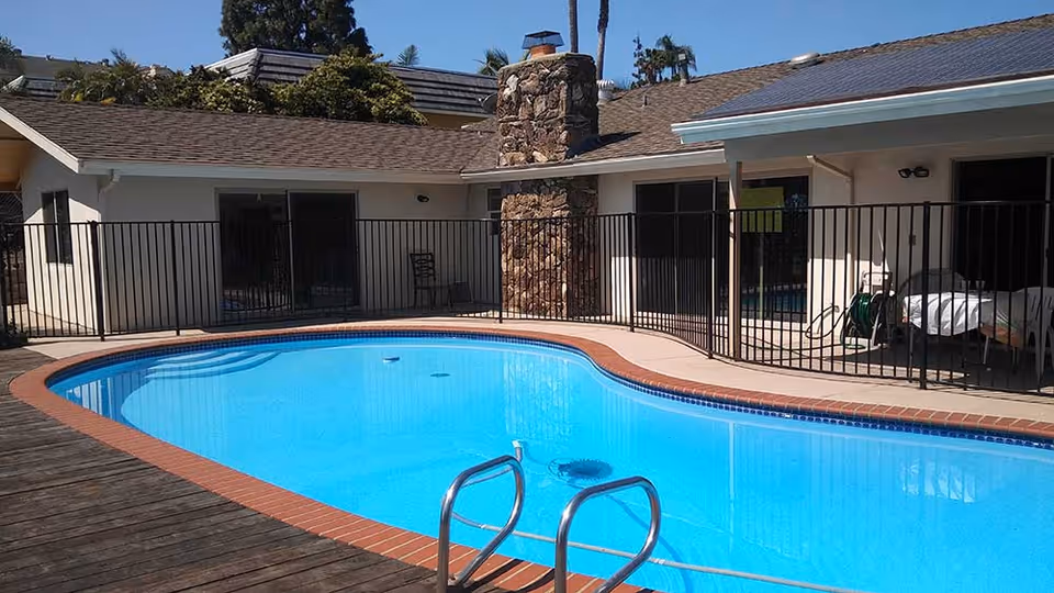 Outdoor swimming pool with clear blue water surrounded by a brick and concrete deck. The pool area is enclosed by a black metal fence. Behind the pool, there is a single-story building with a stone chimney and sliding glass doors. Some outdoor furniture and a garden hose are visible near the building.