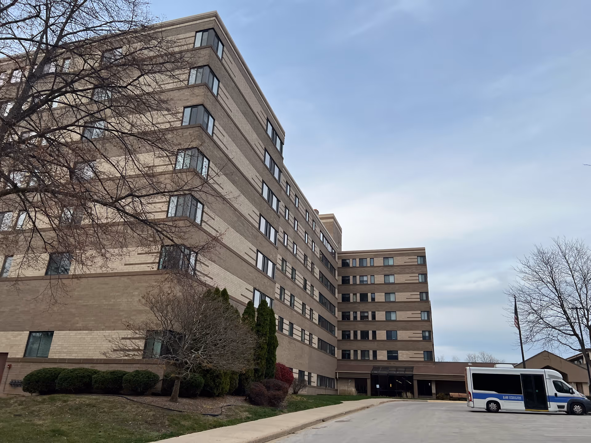 Exterior view of a multi-story senior living facility building with beige and brown brick facade, several windows, a driveway in front, some leafless trees, and a white shuttle bus parked near the entrance under a cloudy sky.