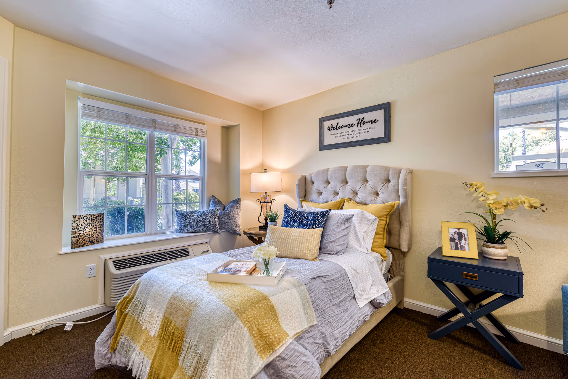 A cozy bedroom with a tufted beige headboard bed dressed in gray and yellow bedding, a yellow and white checkered throw blanket, and multiple pillows. A tray with a book and flowers is on the bed. There is a window with white blinds letting in natural light, a round side table with a lamp and plant, and a blue nightstand with a framed photo and a yellow orchid plant. A framed sign above the bed reads 'Welcome Home'.