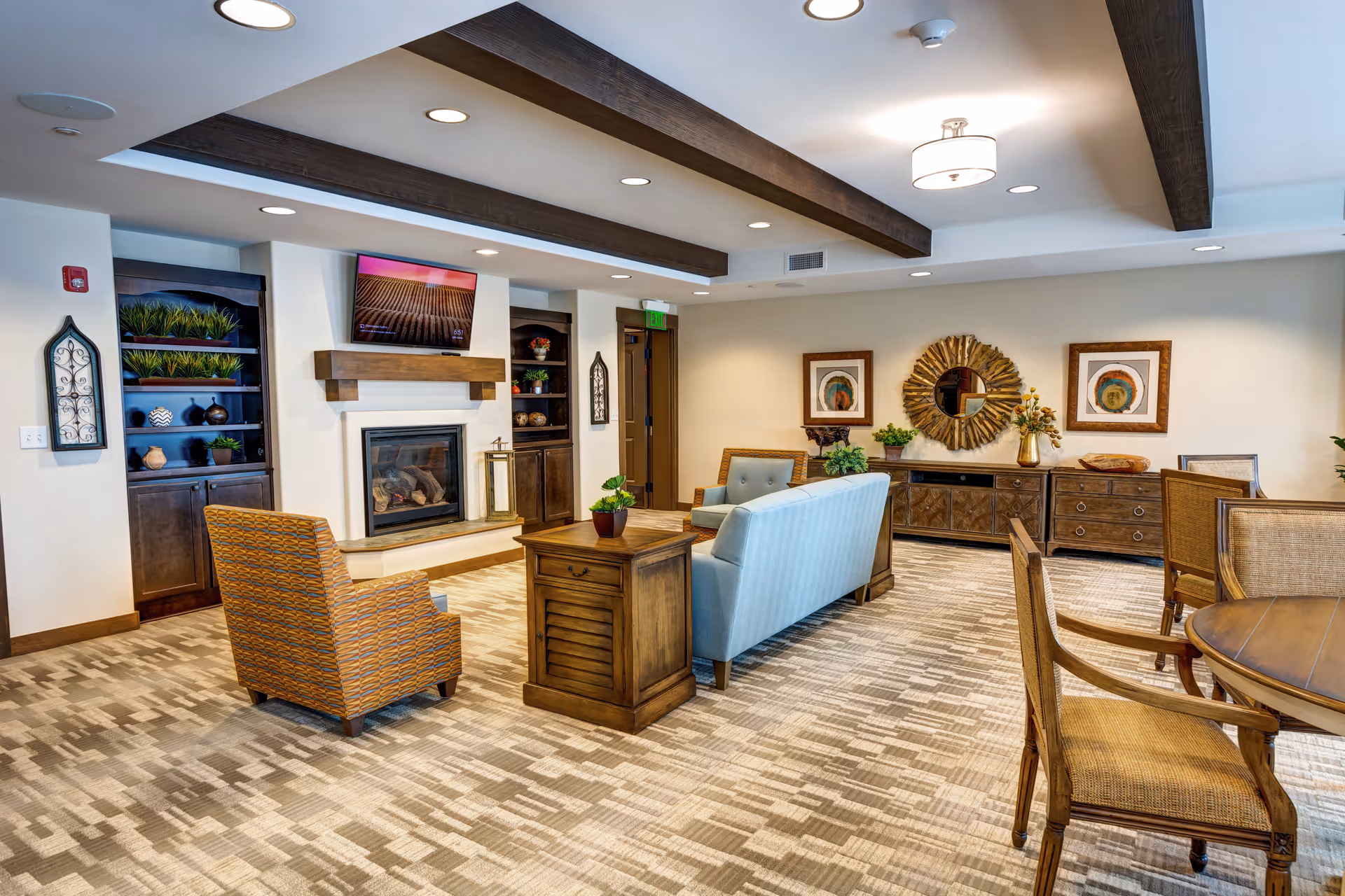 A cozy senior living common area with a light blue sofa, an orange patterned armchair, wooden furniture including a coffee table and sideboard, a fireplace with a mounted TV above it, decorative plants, framed artwork, and ceiling beams with recessed lighting.