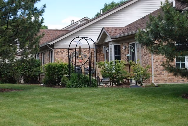 Exterior view of a brick healthcare facility building with a well-maintained green lawn, bushes, and trees. There is a black metal garden arch with a hanging bird feeder in the foreground.