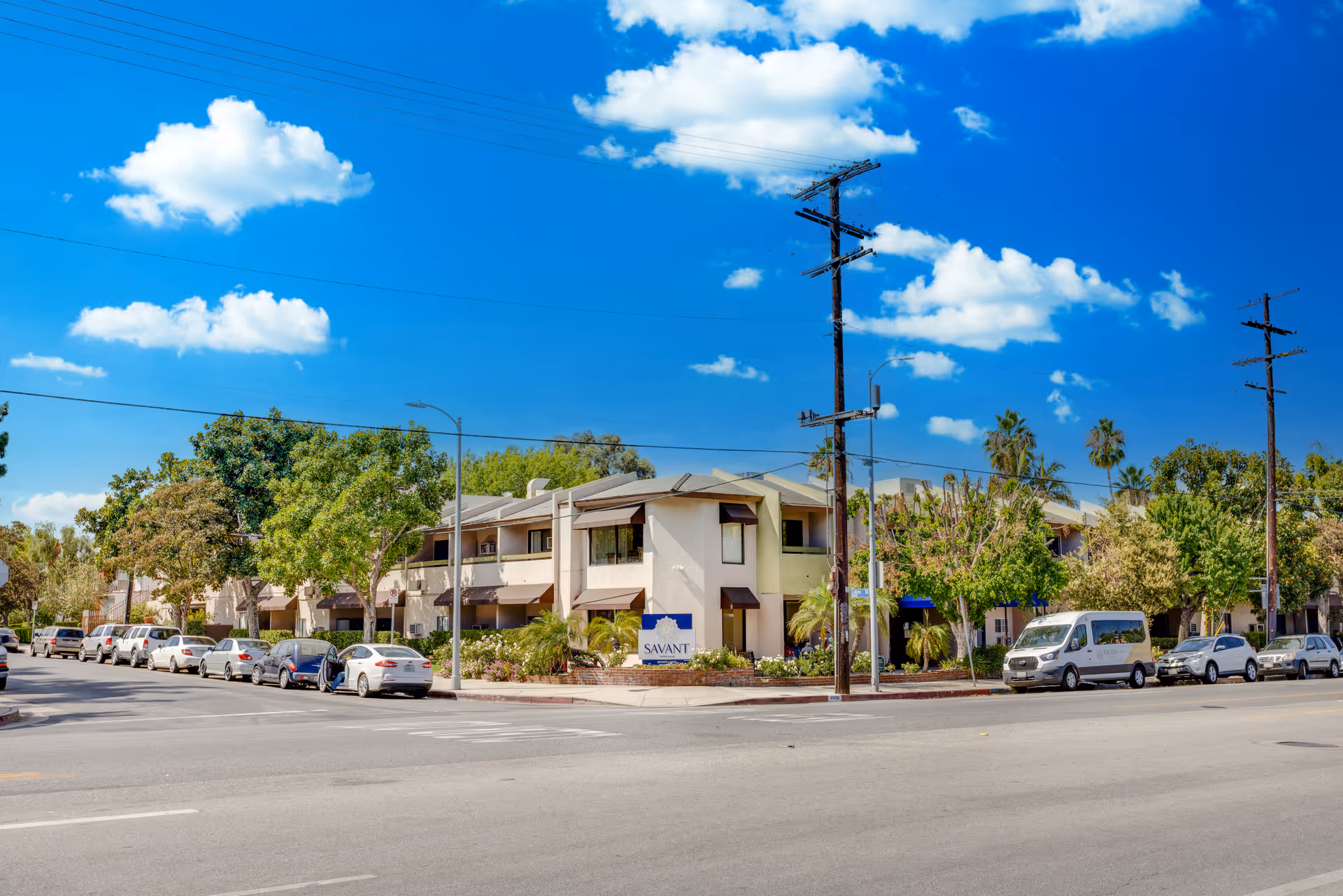 Exterior view of a two-story senior living facility named Savant of Tarzana, surrounded by trees and parked cars along the street under a bright blue sky with scattered clouds.