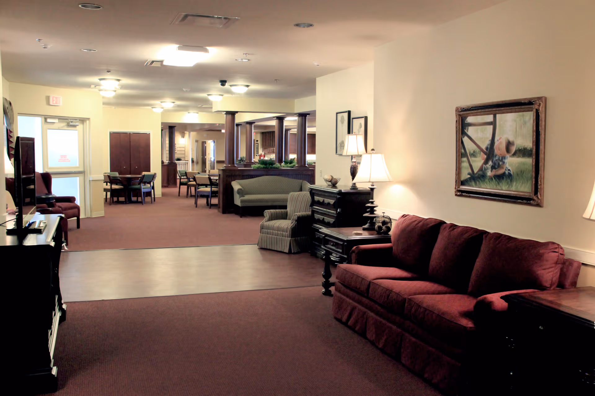 Interior view of a senior living facility lounge area with maroon carpet and a maroon sofa on the right. There are several chairs, tables, and lamps along the walls, with framed artwork hanging. The space extends into a dining area with multiple tables and chairs, and wooden columns separating the areas.
