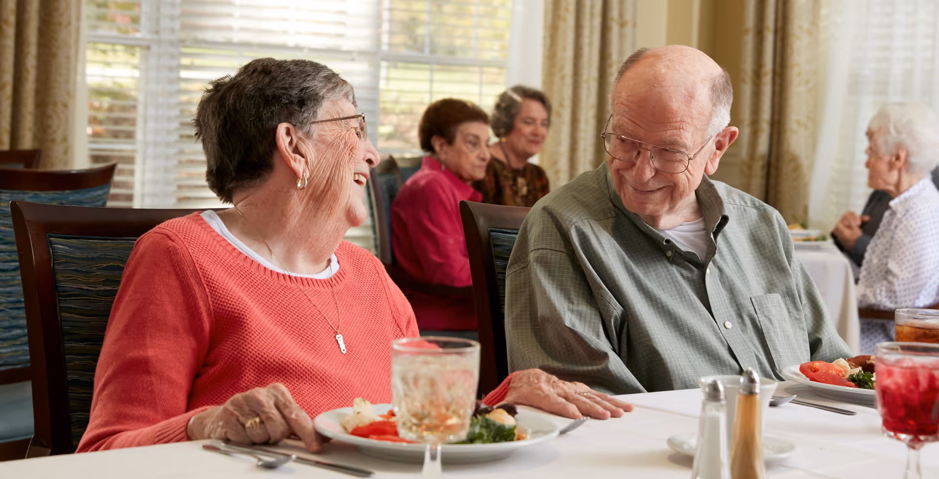 Two elderly residents laugh and share a meal at a communal dining table with other residents seated in the background.