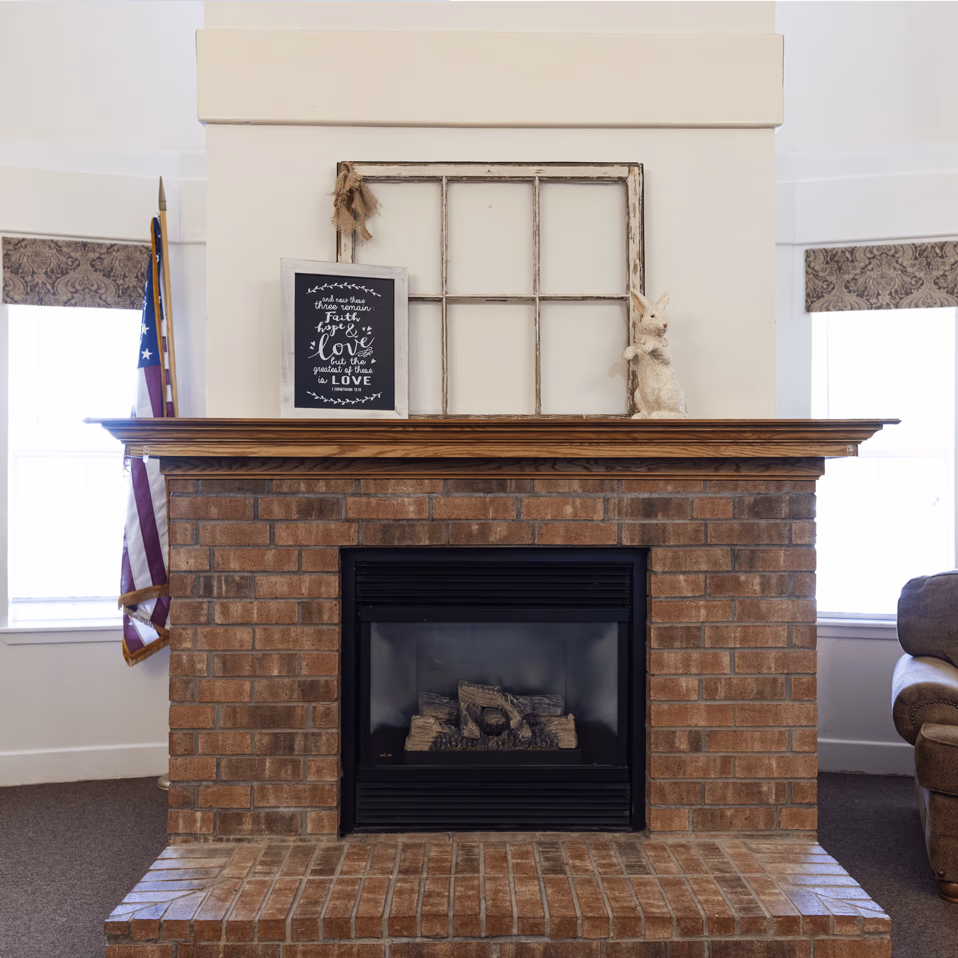 A brick fireplace with a wooden mantel in a living room. On the mantel, there is a rustic window frame, a framed sign with a quote about faith, hope, and love, and a decorative white rabbit figurine. An American flag is visible to the left, and part of a brown upholstered chair is seen on the right. Two windows with patterned valances flank the fireplace.