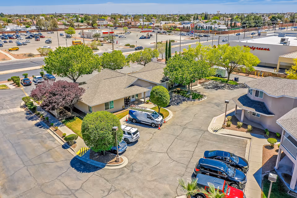 Aerial view of a senior living complex showing single-story buildings, a tree-lined parking lot with parked cars, and nearby commercial stores.