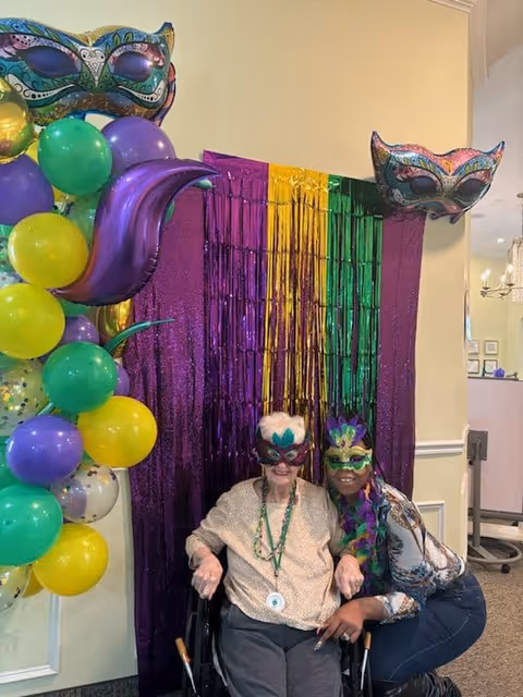Two women wearing colorful Mardi Gras masks posing together in front of a purple, yellow, and green tinsel backdrop. One woman is seated in a wheelchair and the other is crouching beside her. There are balloons in purple, green, and yellow colors on the left side, along with two decorative Mardi Gras masks hanging on the wall.