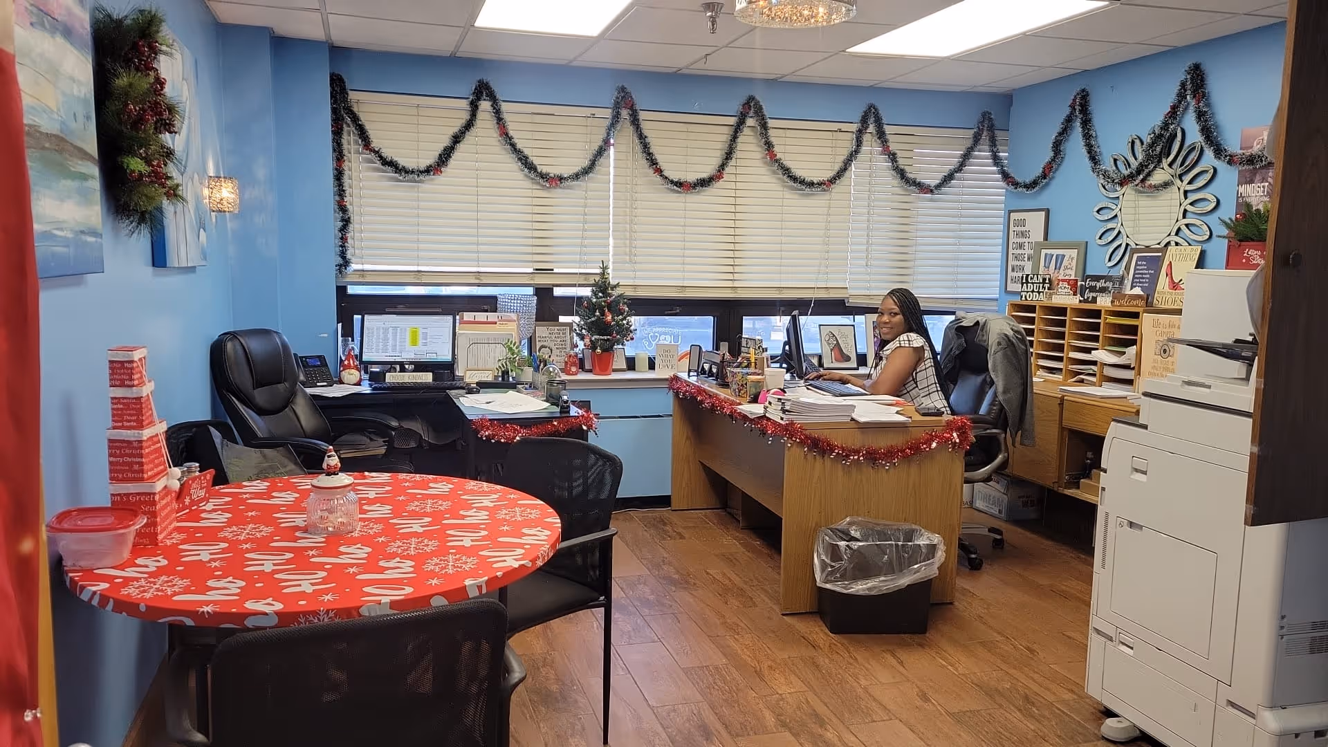 An office room decorated with Christmas garlands and wreaths. A woman is sitting at a wooden desk with a computer, papers, and office supplies. There is a round table covered with a red holiday-themed tablecloth and surrounded by black chairs. The walls are painted light blue, and there are windows with blinds behind the desk. A large printer is visible on the right side of the room.