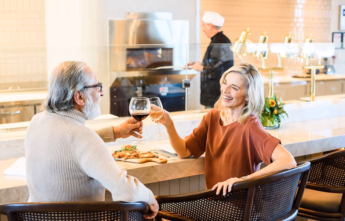 An elderly man and woman sitting at a counter in a dining area, clinking wine glasses and smiling. There is a pizza on the counter in front of them, and a chef in the background preparing food in a kitchen area.