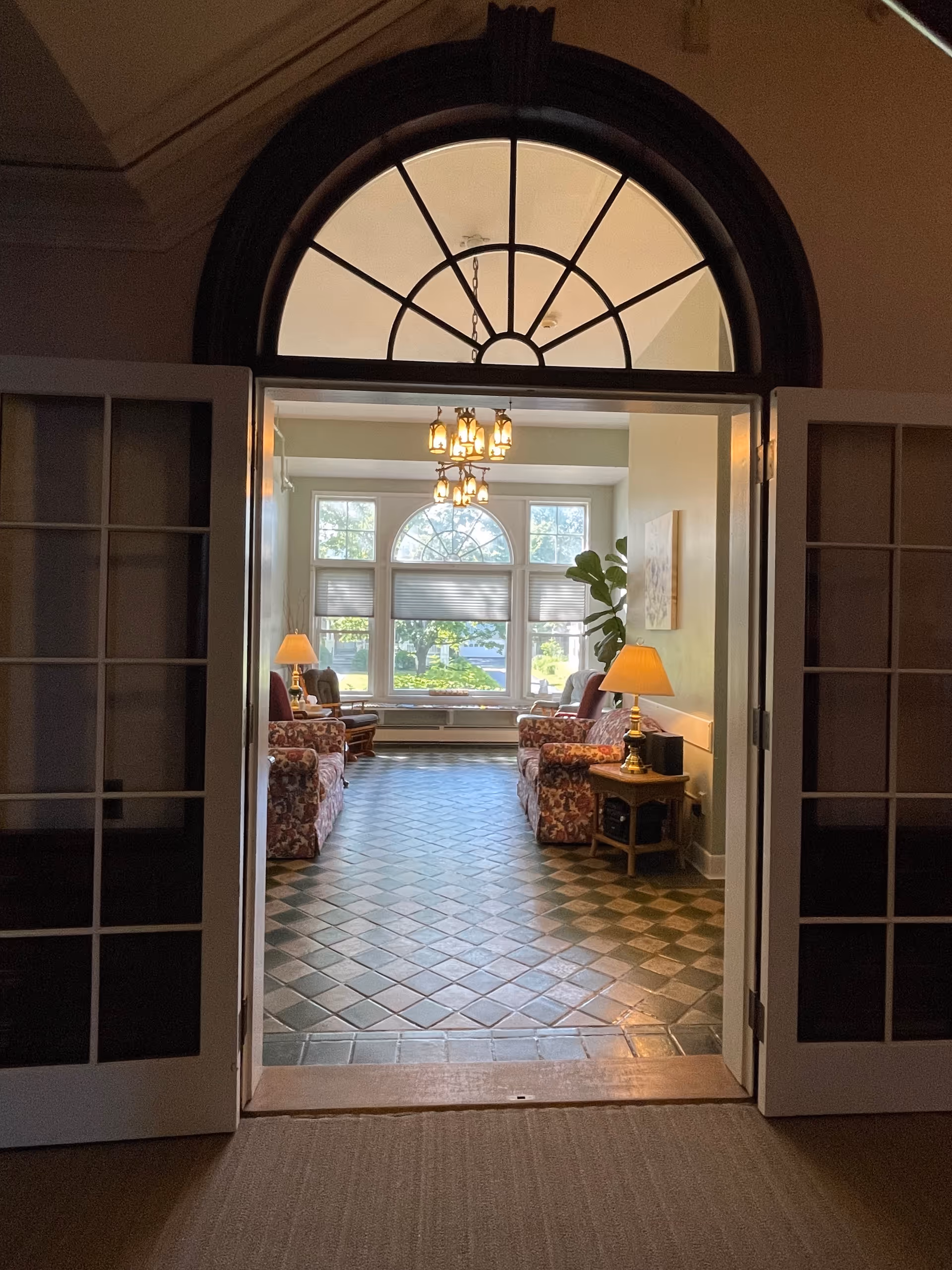 View through open double doors into a sunlit sitting room with patterned armchairs, table lamps, and a large arched window.