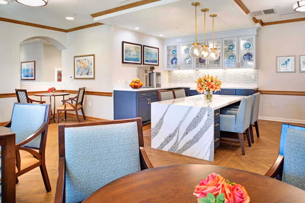 A bright and elegant dining area in a senior living facility featuring a large marble island with blue upholstered chairs, pendant lights hanging above, and a side counter with a coffee machine and fruit bowl. The room has wooden flooring, framed artwork on the walls, and additional seating with wooden chairs and tables, decorated with fresh flowers.