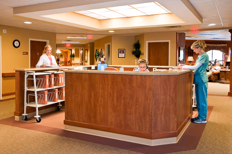 A reception desk area in a senior living facility with two staff members working. One staff member in a white coat is handling files on a cart, while another in green scrubs is writing on a clipboard. The area is well-lit with warm colors and has a hallway and seating area visible in the background.
