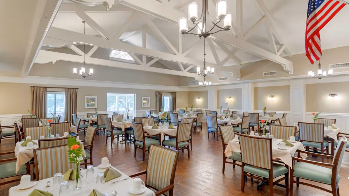 Large dining room with multiple set tables and striped chairs under white exposed trusses and chandeliers, an American flag hanging in one corner.